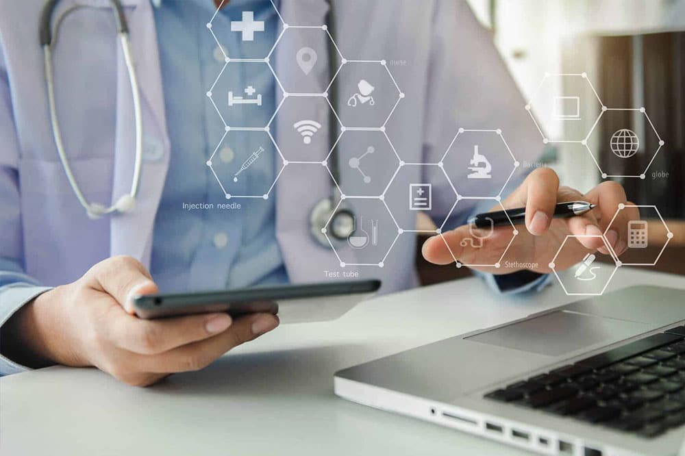 Healthcare physician sitting at a desk with tablet entering in data to laptop.